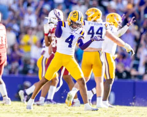 LSU Tigers 2026 NFL Draft prospect wearing a white No. 4 jersey celebrates on the field with an emphatic pose after a big defensive play, with teammates and a packed stadium in the background.