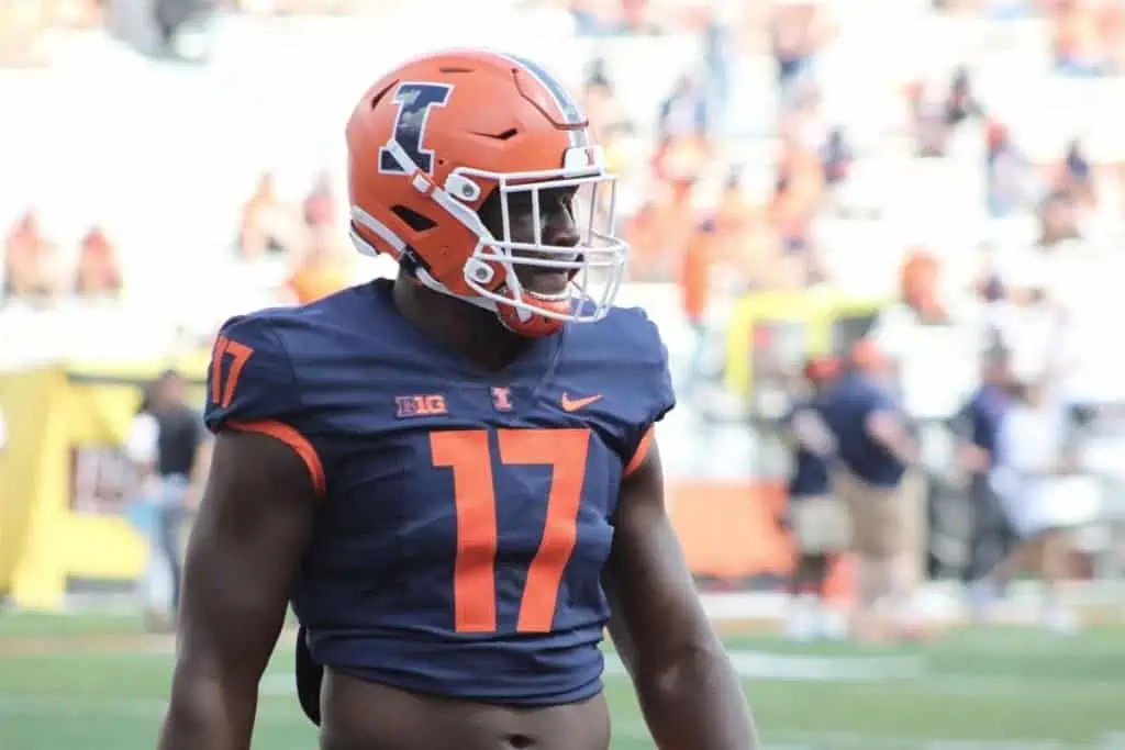 Illinois football player, Gabe Jacas, in a navy jersey and orange helmet, number 17, walks on the field during pregame warmups.
