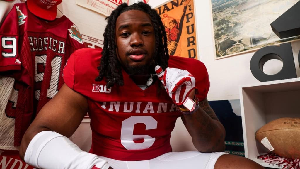 Indiana Hoosiers football player Mikail Kamara, wearing a red No. 6 uniform, sits indoors and points toward the camera with memorabilia and team posters visible in the background.