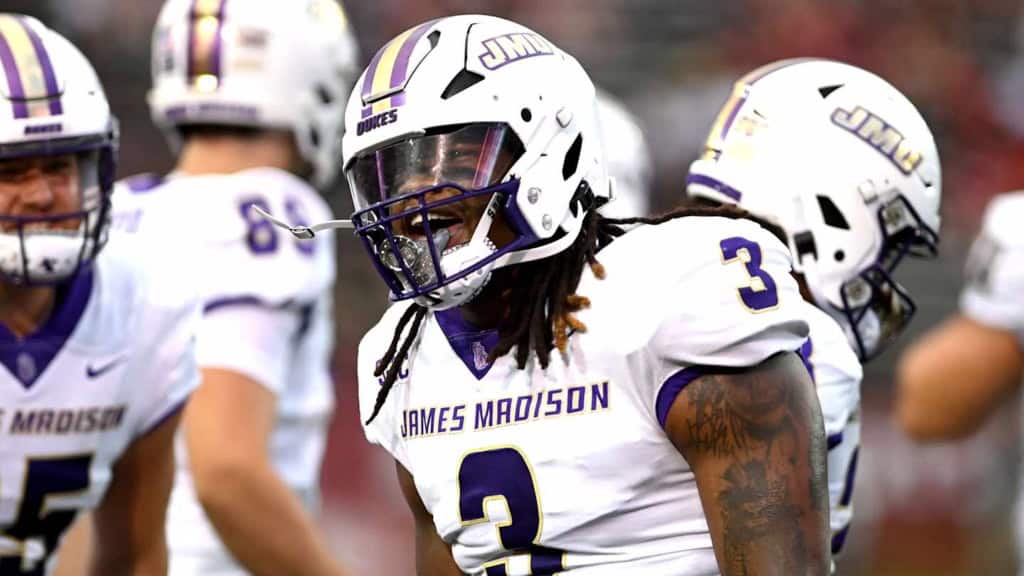 James Madison Dukes defensive player Mikail Kamara, wearing a white No. 3 uniform and helmet, stands on the field during a game with teammates lined up behind him.