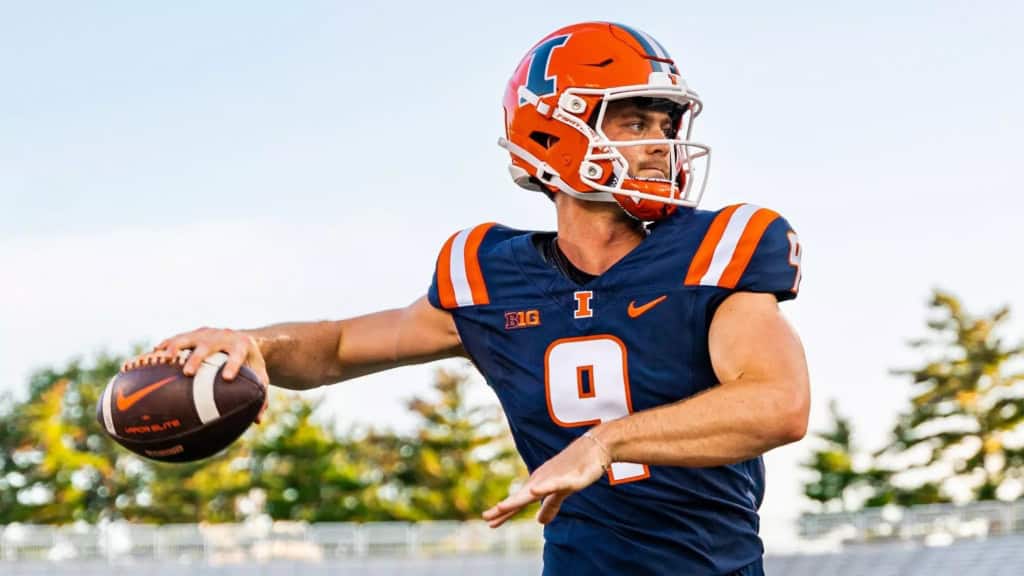 Illinois quarterback #9 drops back to pass during a game, wearing an orange helmet and navy uniform while gripping the football in throwing motion.