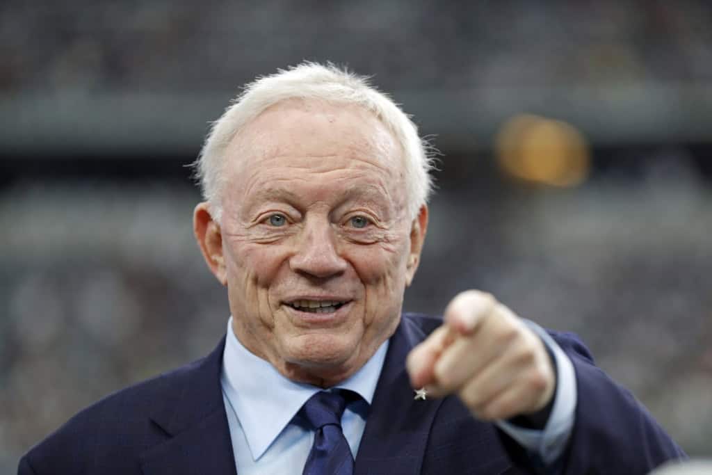 Close-up of Jerry Jones in a suit and tie pointing directly toward the camera on the field at AT&T Stadium.