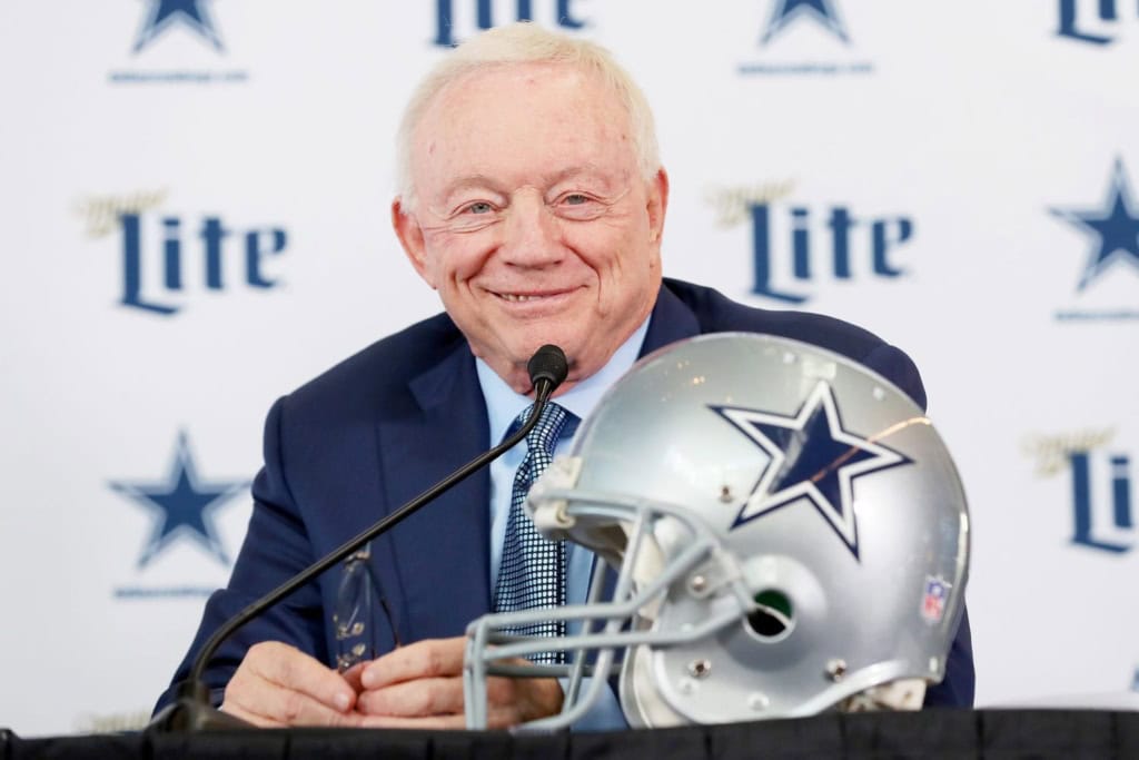 Jerry Jones seated at a press conference table with a Dallas Cowboys helmet in front of him, speaking into a microphone with a backdrop featuring Cowboys star logos.
