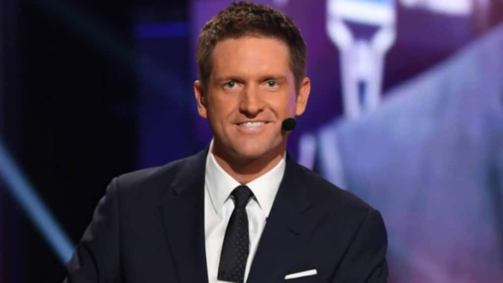 Todd McShay on stage during NFL Draft broadcast, dressed in a suit with microphone headset while delivering draft analysis.
