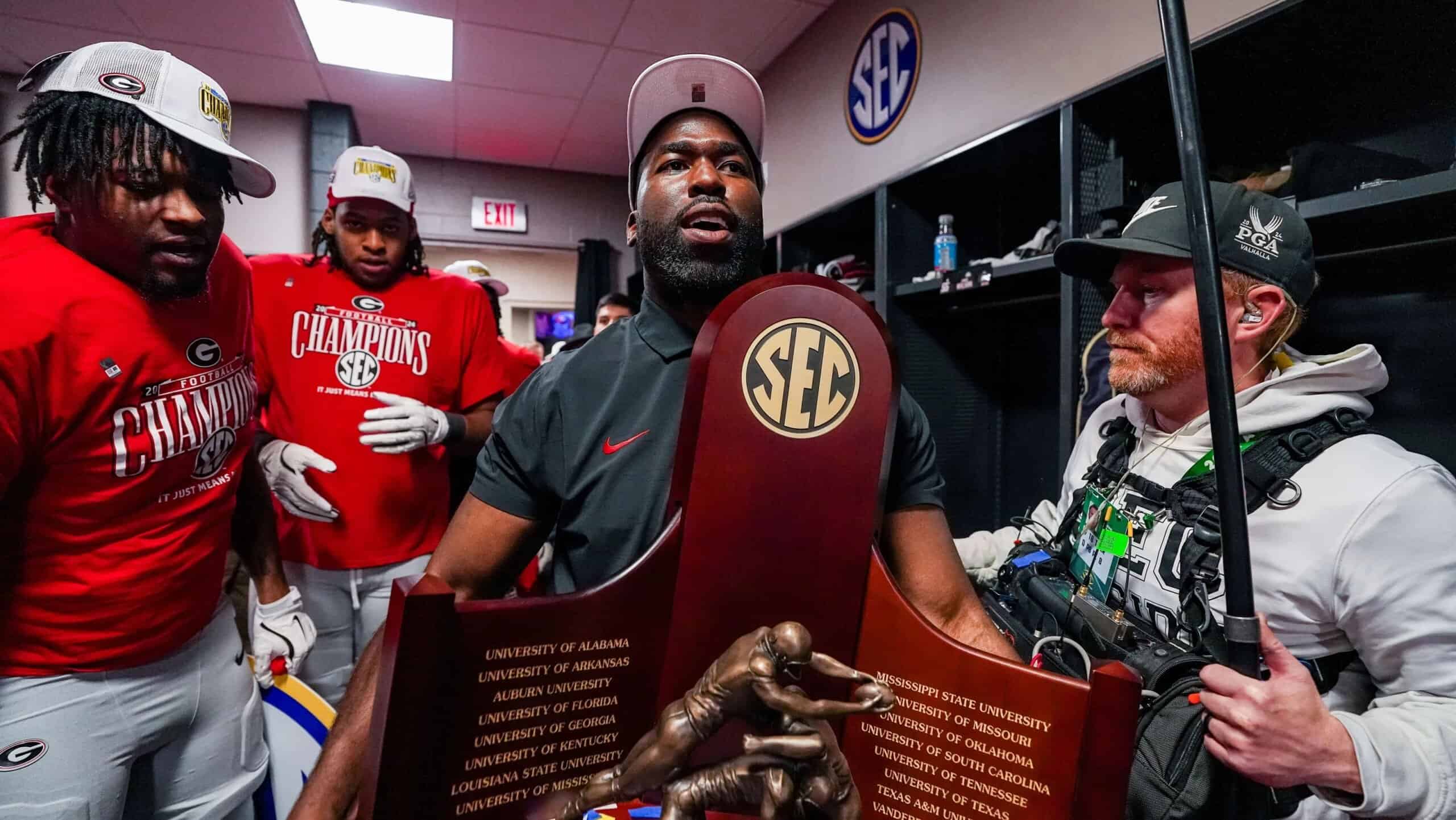 Georgia Bulldogs defensive coach celebrating with players in the locker room while holding the SEC Championship trophy after the game.