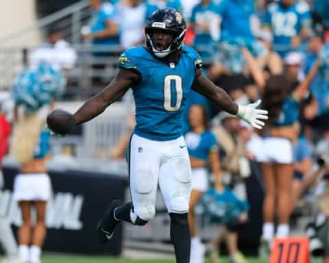 Devin Lloyd celebrates during a Jacksonville Jaguars game, holding the football in his right hand while spreading his arms wide in a teal number 0 jersey as the crowd looks on in the background.