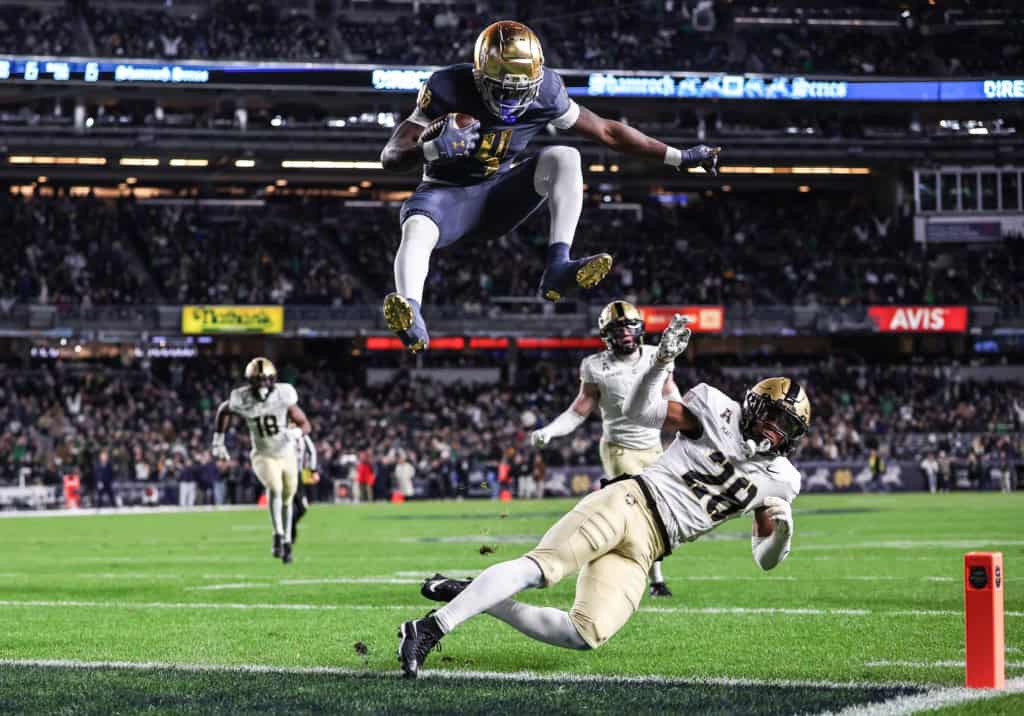 Notre Dame running back Jeremiyah Love leaps over a defender near the goal line during a night game, showing explosive athleticism and balance.