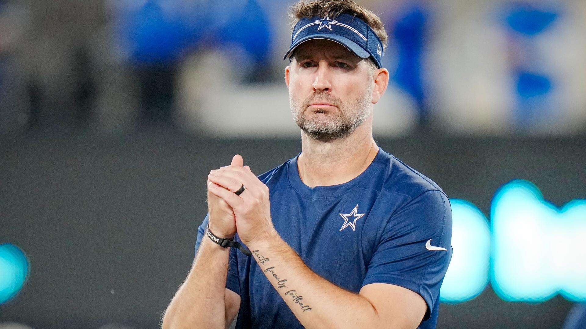 Dallas Cowboys head coach Brian Schottenheimer stands on the sideline during warmups, wearing a navy visor and blue team shirt with a focused expression.