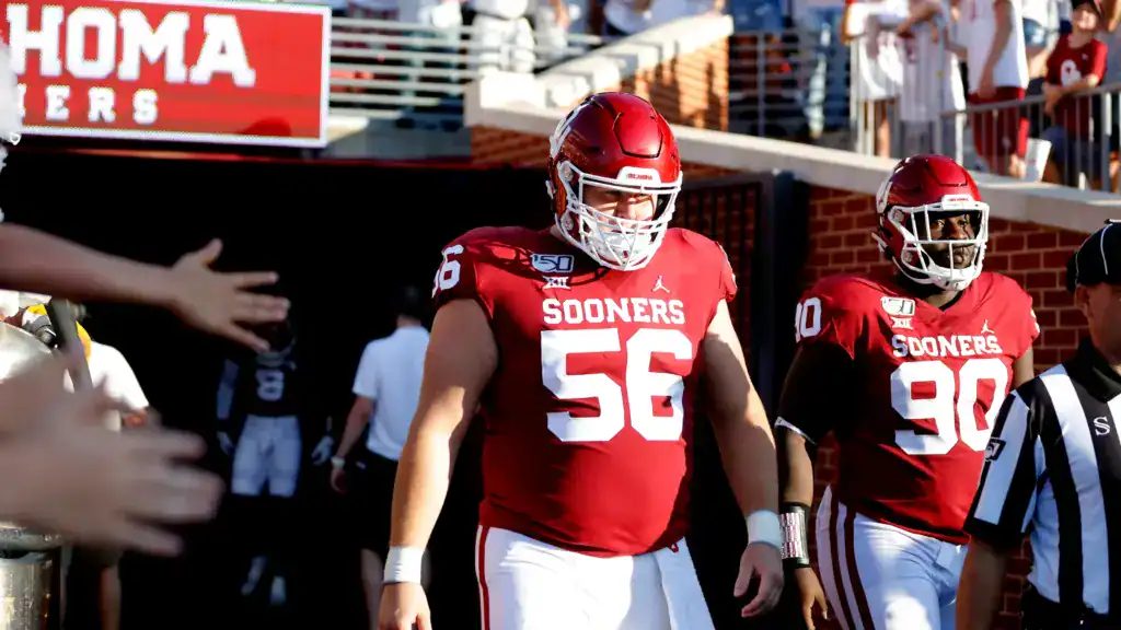 Oklahoma Sooners linemen walk out of the tunnel in a college football game, representing university draft picks that earned First Team All-Pro honors in the NFL.
