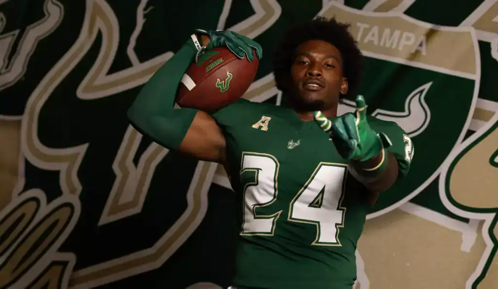 2026 NFL Draft prospect Mac Harris of USF poses in a green Bulls uniform during a photo shoot, holding a football on his shoulder in front of a USF backdrop.