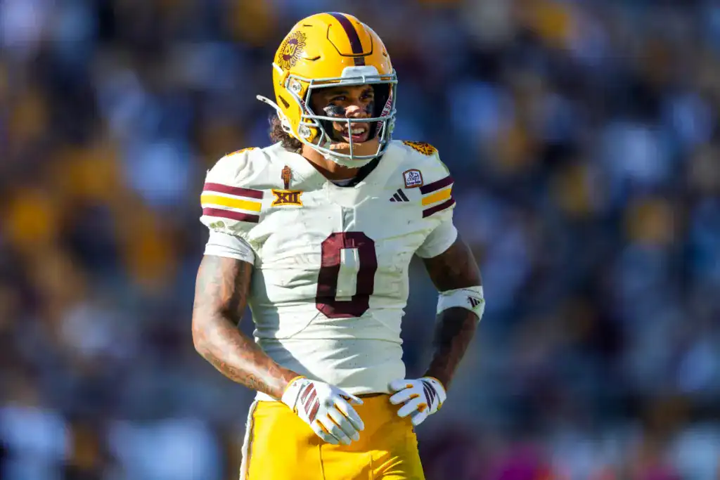 Jordyn Tyson stands on the field in Arizona State’s white road uniform and gold helmet, smiling with his hands near his waist during a game. 