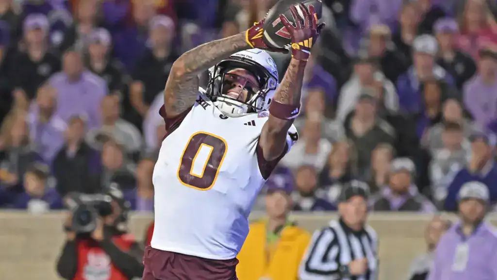 Jordyn Tyson reaches up to make a catch in Arizona State’s white uniform during a game, with a blurred crowd in the background.