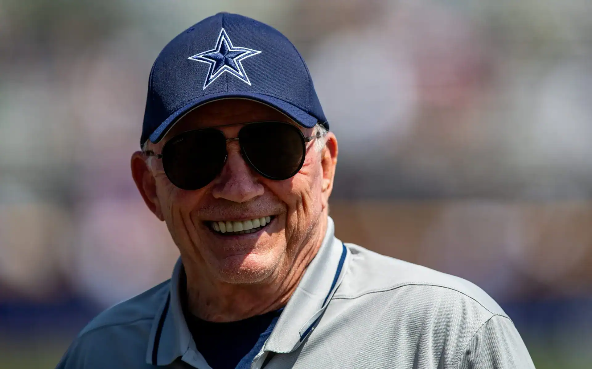 Dallas Cowboys owner Jerry Jones smiles while wearing a Cowboys cap and sunglasses on the sideline.