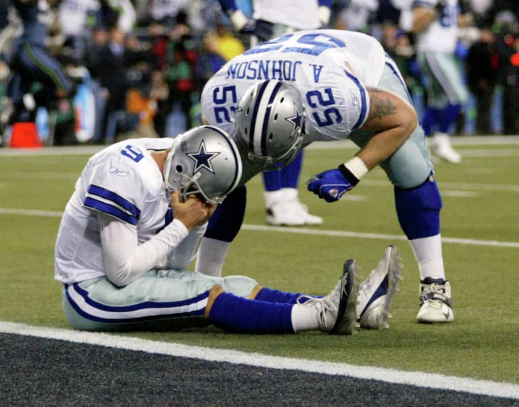Dallas Cowboys quarterback Tony Romo sits on the field as center Al Johnson checks on him in a historic image featured in a Cowboys preseason history story.