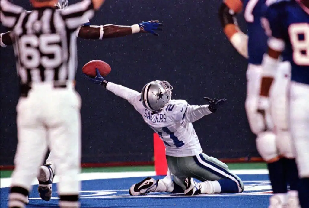 Deion Sanders stretches the football out in one hand while celebrating on the field for the Dallas Cowboys.