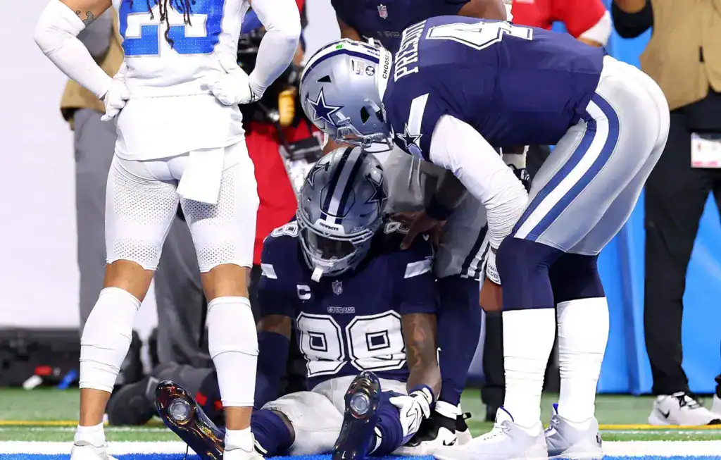 CeeDee Lamb sits on the field while Dallas Cowboys teammates gather around him following a play.