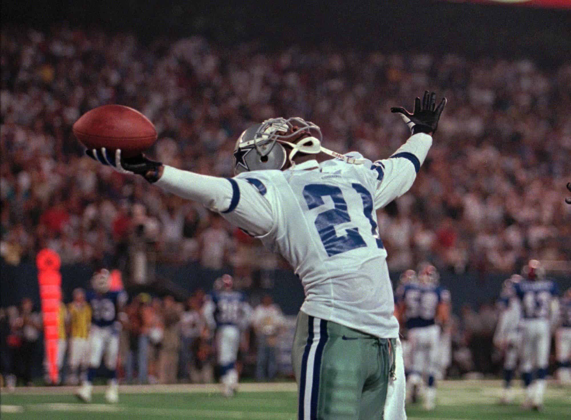 Deion Sanders celebrates while holding the football high in one hand during a Dallas Cowboys game.