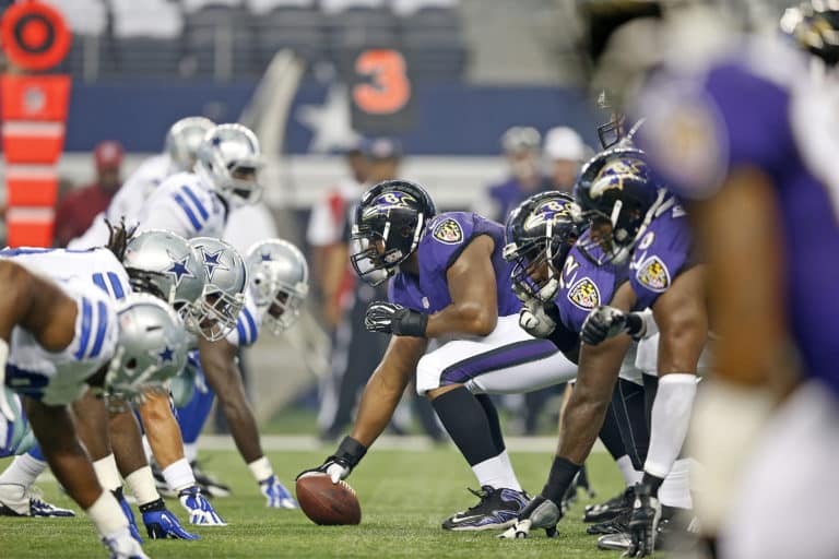 American football teams in purple and white uniforms are lined up facing each other before the snap.