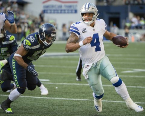 A football player in a white and blue uniform runs with the ball while being chased by an opponent in a blue and green uniform.