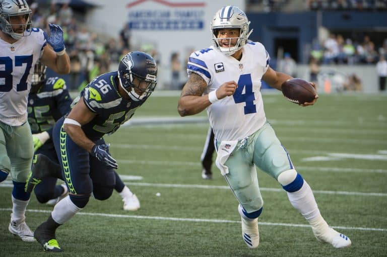 A football player in a white and blue uniform runs with the ball while being chased by an opponent in a blue and green uniform.