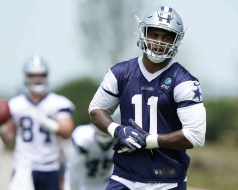 A football player in a navy and white uniform practices at Training Camp, with a teammate holding a football in the background.