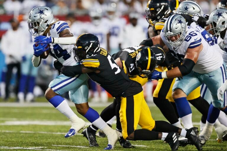 Football players from opposing teams in action on the field, one player carrying the ball while others attempt a tackle.