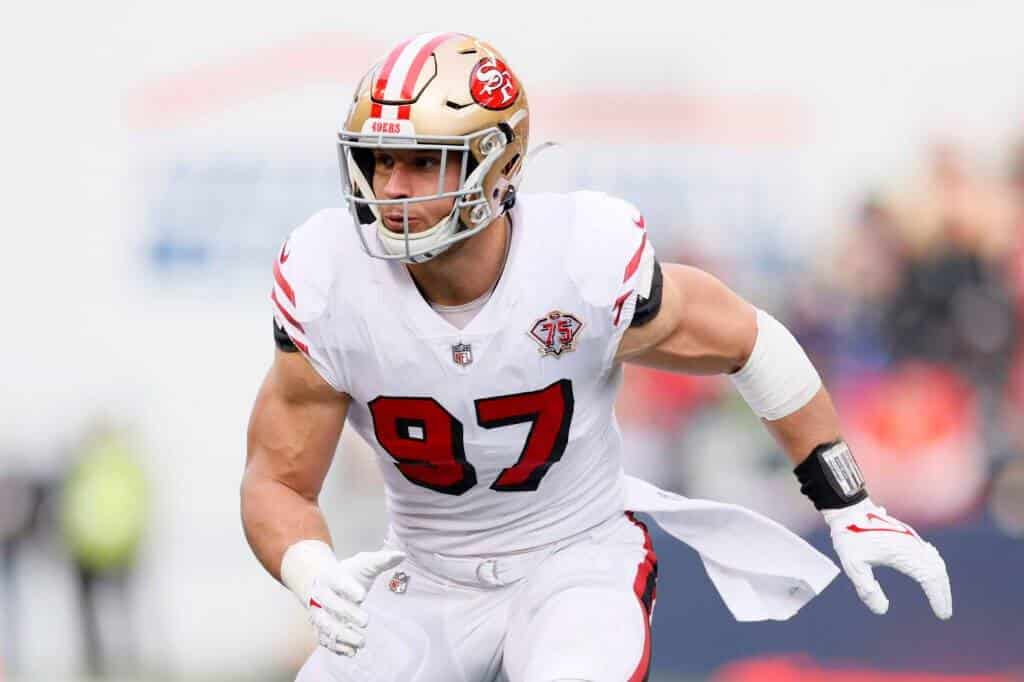 Football player Nick Bosa runs across the field during a game wearing a white 49ers uniform with the number 97 and a gold helmet.