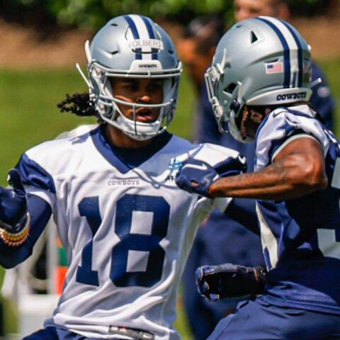 Two football players in Dallas Cowboys uniforms and helmets, including Jalen Tolbert, practice on the field with teammates in the background.
