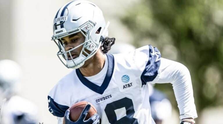 Football player in a white and blue uniform running with the ball during practice, wearing a helmet bearing the name "Jalen Tolbert," representing the Cowboys.