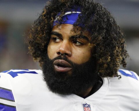A football player with a beard and curly hair, resembling Ezekiel Elliott, wearing a white and blue jersey glances to the side on the field during preseason play.