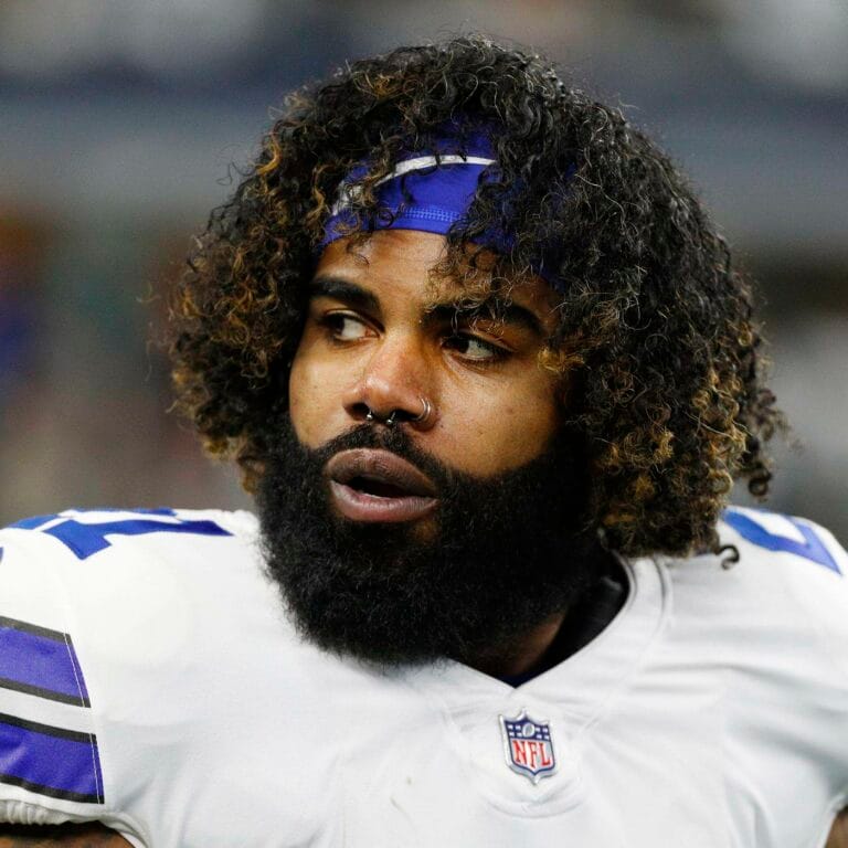 A football player with a beard and curly hair, resembling Ezekiel Elliott, wearing a white and blue jersey glances to the side on the field during preseason play.