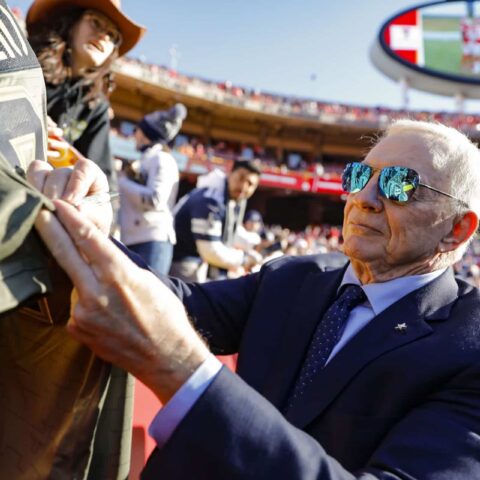 An elderly man in a suit and sunglasses signs a jacket at a sports stadium.