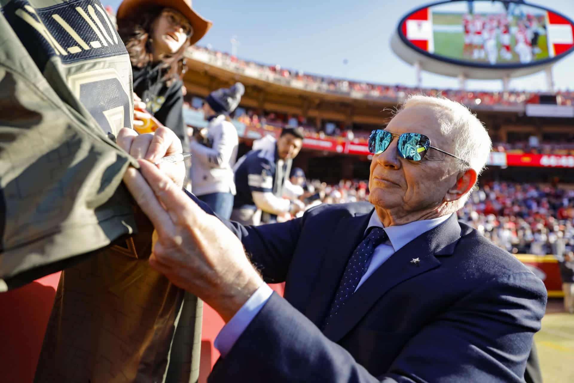 An elderly man in a suit and sunglasses signs a jacket at a sports stadium.