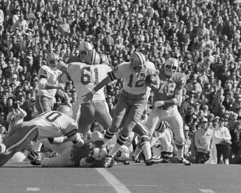 Black and white photo of a football game with players from both teams in action, featuring the Cowboys' legendary Roger Staubach. A crowd of spectators can be seen in the background.