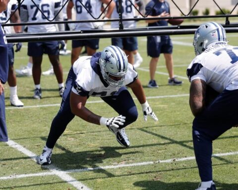Football players in jerseys and helmets practice on a grassy field, with coaches observing them closely. Among them, Chuma Edoga stands out as he hones his skills with precision and determination.