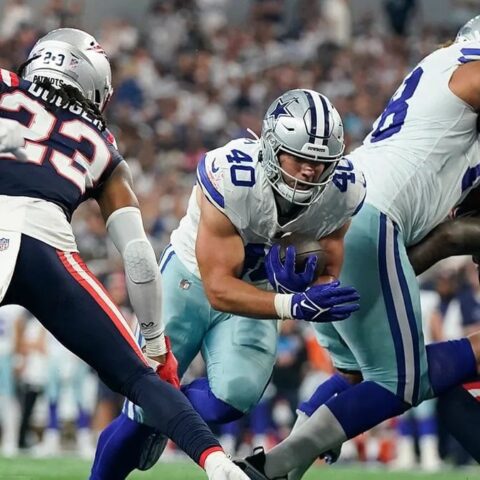 Football players from opposing teams in action during a game, with one team in blue and white uniforms and the other in blue and red. Hunter Luepke of the Cowboys makes a pivotal play that has fans on their feet.