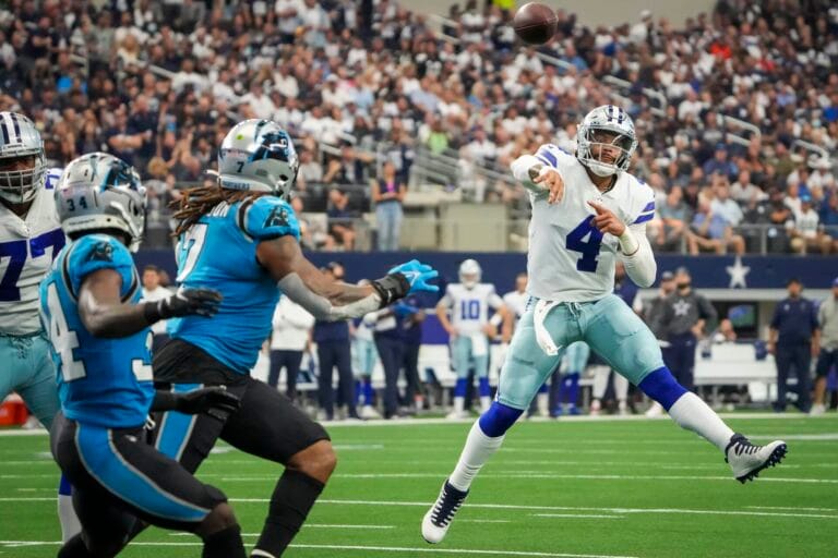 A football player in a white uniform, resembling Dak Prescott, throws a pass while three Panthers players in blue and black attempt to block the throw.