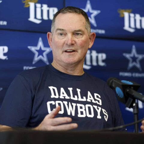 A man wearing a Dallas Cowboys shirt speaks at a press conference with a backdrop of Cowboys logos and Miller Lite branding.