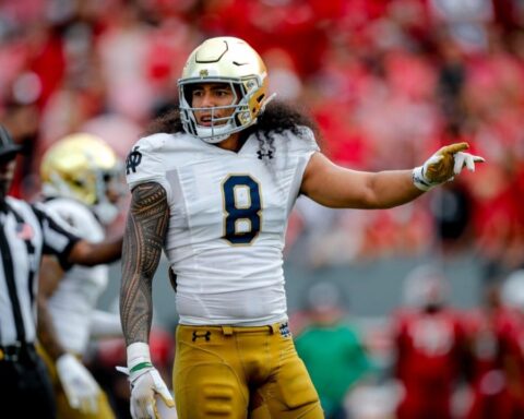 A football player in a white and yellow uniform with number 8, resembling Marist Liufau, stands on the field during a game, gesturing with his left hand.