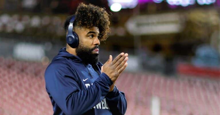 A man with headphones and curly hair, resembling Ezekiel Elliott in his focus, wears a dark blue hoodie as he stands with hands clasped in an outdoor setting.