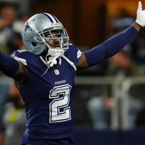 Football player in a blue jersey and helmet raises arms while holding a football on the field.
