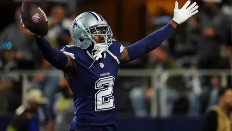 Football player in a blue jersey and helmet raises arms while holding a football on the field.