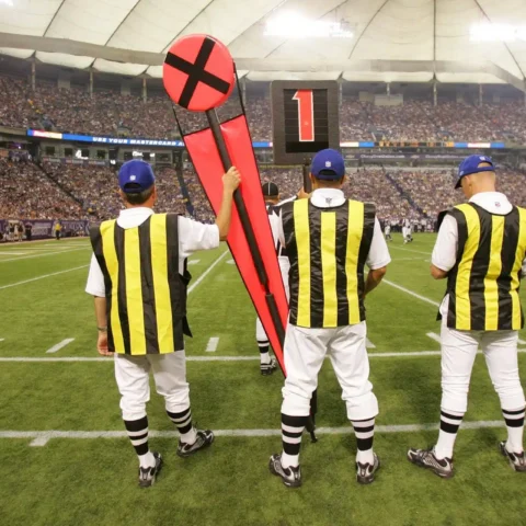 Three officials in striped uniforms hold yardage markers on the sidelines during a football game in a stadium, seamlessly integrating technology to ensure precise measurements and smooth gameplay.