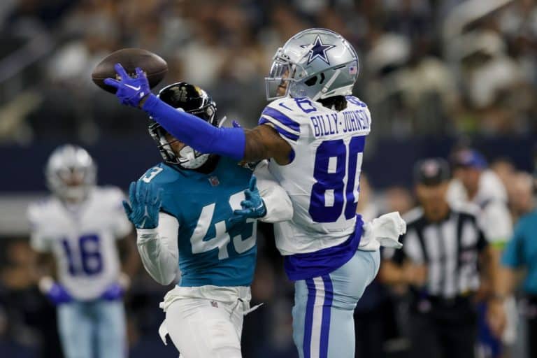 A football player in a blue uniform attempts to catch a pass, while Tyron Billy-Johnson, in a white and blue uniform, tries to block him.