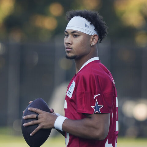 A football player in a red jersey holds a football, standing outdoors with a focused expression.