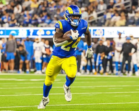 During Cowboys Training Camp, an RB in blue and yellow uniform runs with the ball during a game on a field with a crowd in the background, earning praise for his agility and performance.