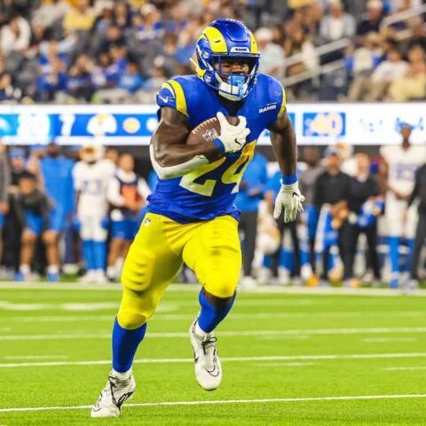 During Cowboys Training Camp, an RB in blue and yellow uniform runs with the ball during a game on a field with a crowd in the background, earning praise for his agility and performance.