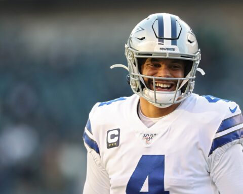 A football player in a white Dallas Cowboys jersey and helmet smiles on the field.