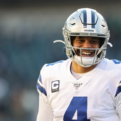 A football player in a white Dallas Cowboys jersey and helmet smiles on the field.