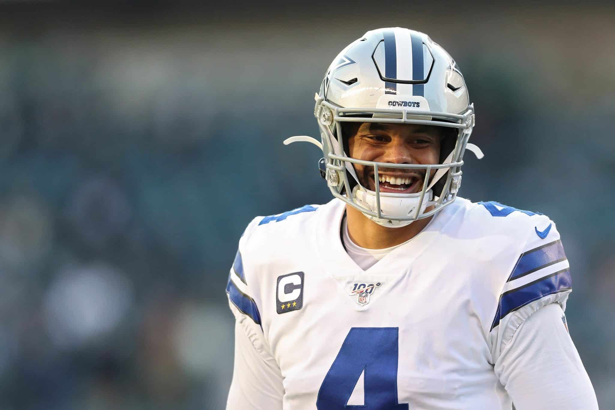 A football player in a white Dallas Cowboys jersey and helmet smiles on the field.